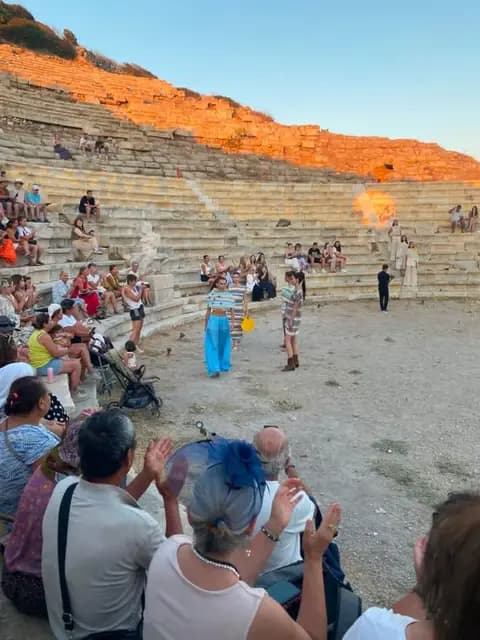 Knidos antik tiyatrosunda seyirci alkışlarken mankenler orkestra alanında yürüyor; üst sıralar gün batımıyla turuncuya boyanmış.