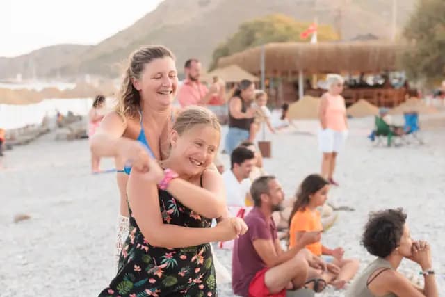 Sahil çakılında iki kadın birbirine sarılıp gülerek dans ederken arkalarındaki izleyiciler onları seyrediyor.
