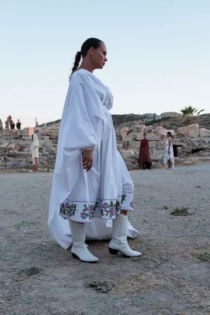 Beyaz işlemeli uzun elbiseyle manken Knidos'un toprak podyumunda yürüyor.
