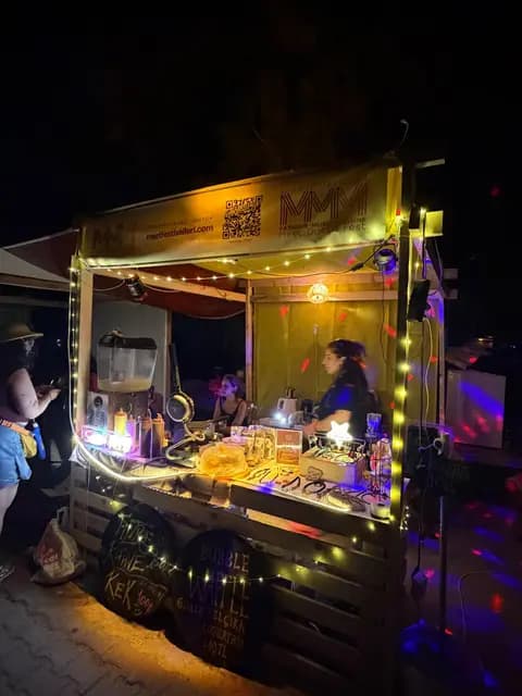 Gece MMM logolu ahşap büfe peri ışıklarıyla parlıyor; içeride görevli sipariş hazırlıyor.