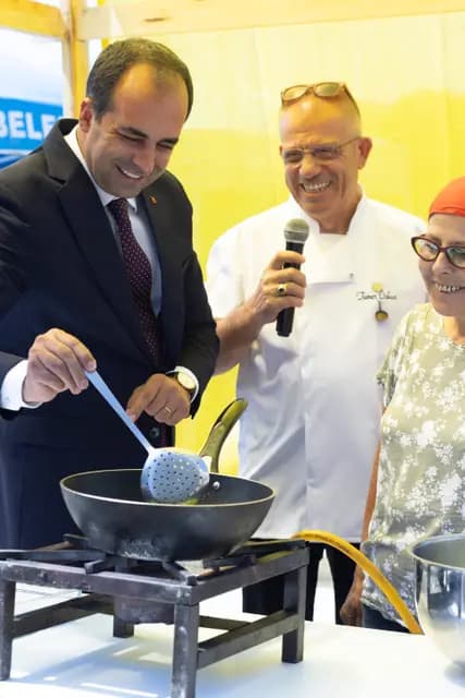 Protokol üyesi ocakta tavaya kepçe daldırırken şef mikrofonla anlatıyor, yanındaki kadın üretici içten gülüyor.