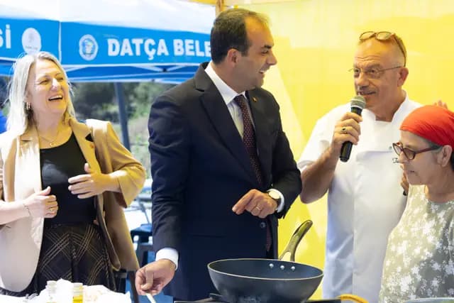 Mutfak gösterisinde şef mikrofonla anlatırken protokol ve davetliler tavaya eğilip gülüyor.