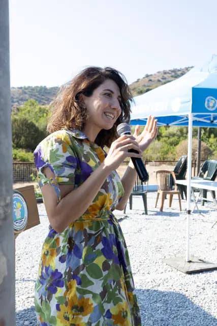 Çiçekli elbiseli kadın konuşmacı elini kaldırarak mikrofonla topluluğa sesleniyor.