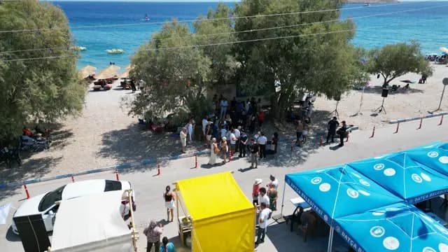 Yüksekten çekilmiş genel kare: turkuaz koy, çam gölgesindeki festival alanı ve belediye şemsiyeleri bir arada.