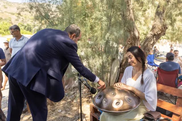 Protokol üyesi eğilip handpan'e dokunuyor; genç kadın müzisyen gülümseyerek izliyor.