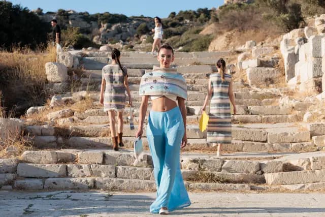 Antik basamaklardan aşağı yürüyen manken öne çıkarken diğerleri arkada podyumu sürdürüyor — Knidos sahnesi tam görünüyor.