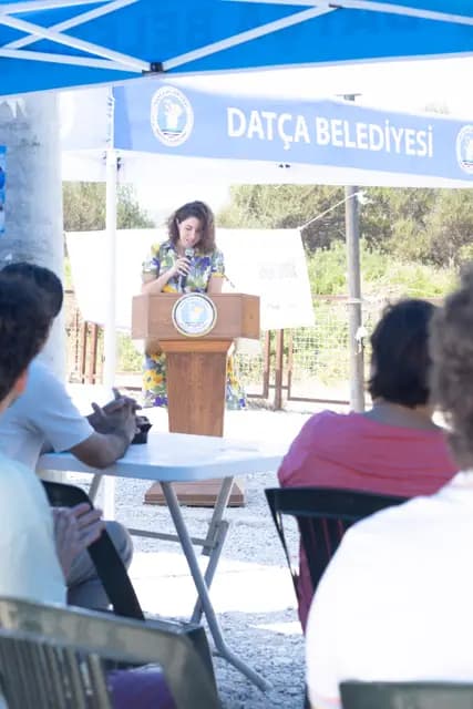 Günün sunucusu kürsüde programı açıyor; arkada Datça Belediyesi gölgeliği.
