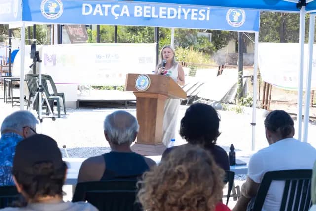 Kürsüdeki konuşmacı dinleyicilere sesleniyor; ön sırada oturanlar not alıyor.