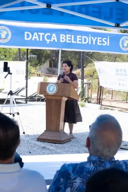 Kürsüde konuşmacı, arkasında festival afişi ve belediye gölgeliğiyle.