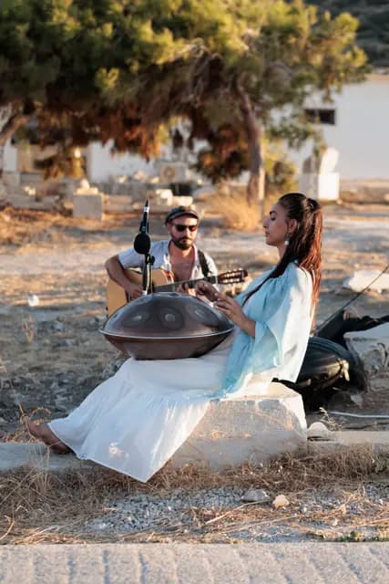 Knidos'ta taşların üzerine oturan müzisyen handpan çalıyor, arkasında koy görünüyor.