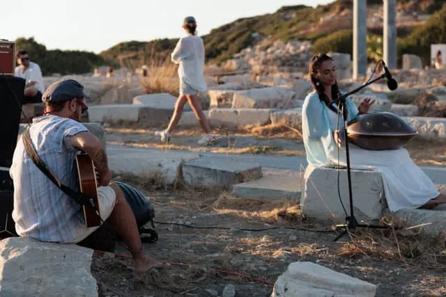 Antik alanda gitar ve handpan birlikte çalınıyor; ziyaretçiler arkadan geçiyor.